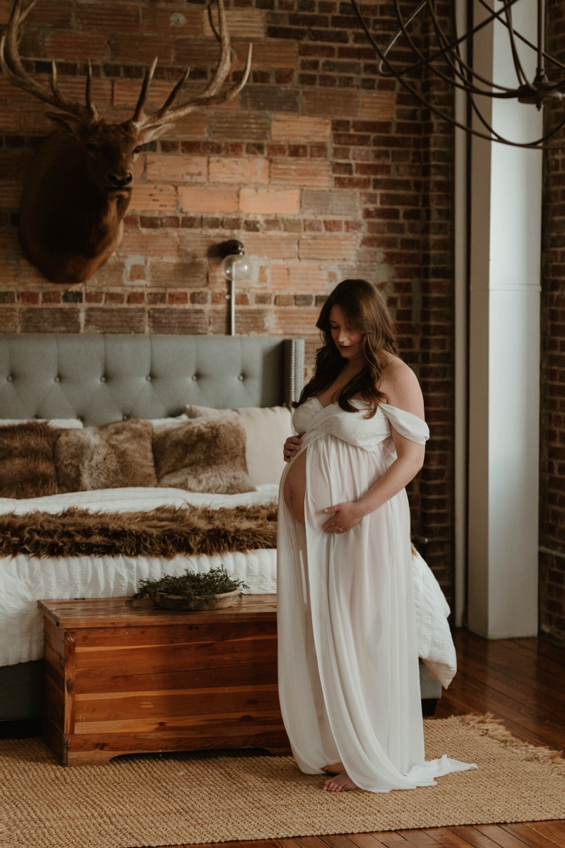 Expectant mother in white dress by exposed brick window